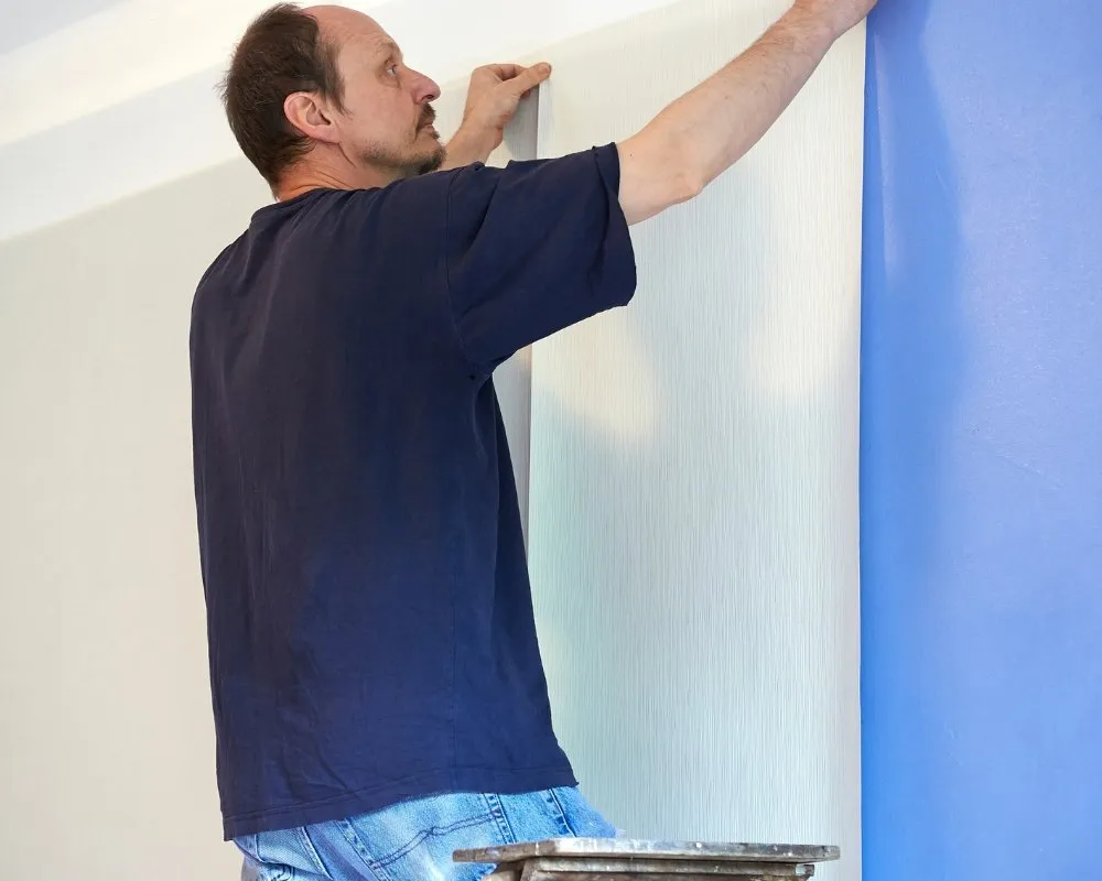 Homem em cima de uma escada alinhando uma tira vertical de papel de parede texturizado ao lado de uma parede azul